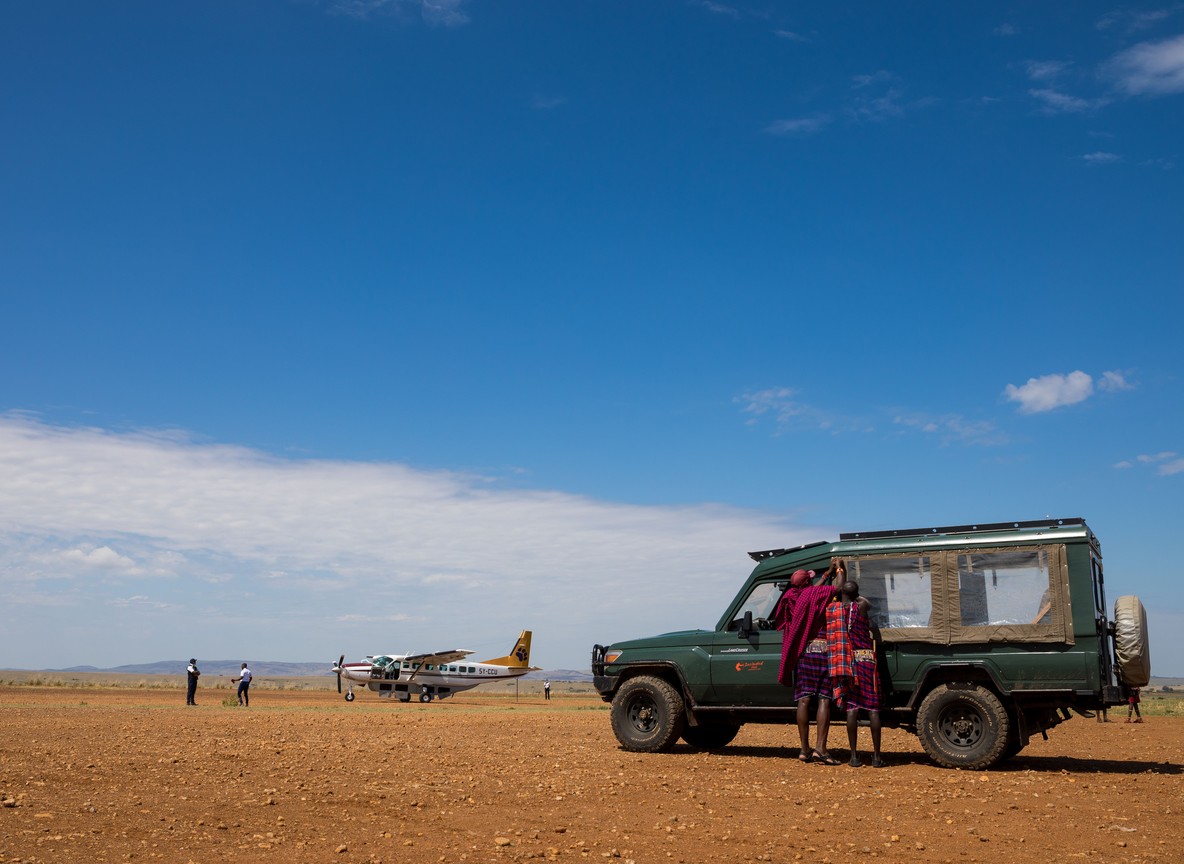 Small bush plane on a red-earth airstrip with an open safari vehicle and Maasai guides waiting nearby, Kenya