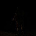 A safari askari in uniform holding a torch and walking a guest along a lit path at an unfenced tented camp at night