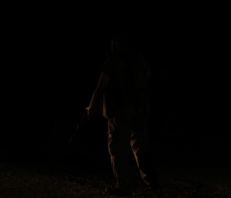 A safari askari in uniform holding a torch and walking a guest along a lit path at an unfenced tented camp at night