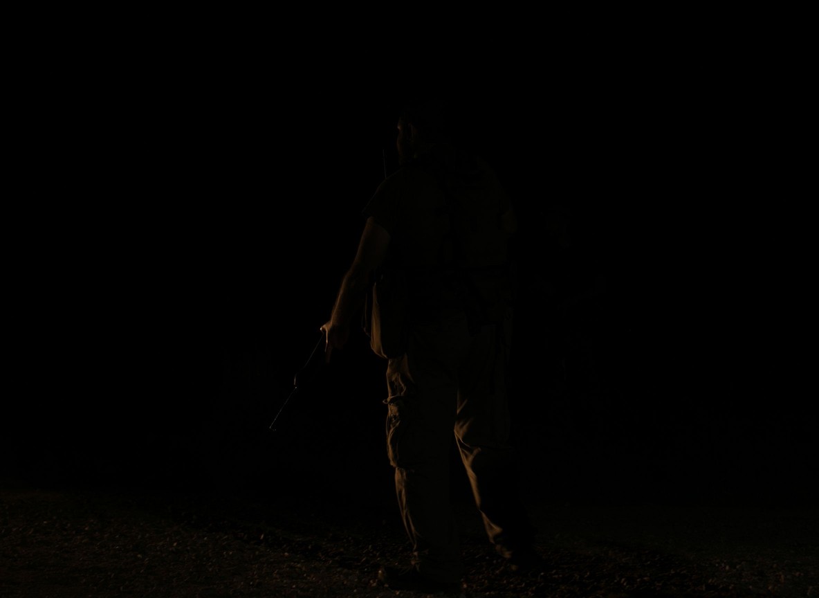 A safari askari in uniform holding a torch and walking a guest along a lit path at an unfenced tented camp at night