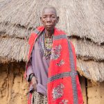 a Maasai elder and a visitor talking directly at the entrance to a manyatta homestead, warm afternoon light, Maasai Mara region grassland behind them