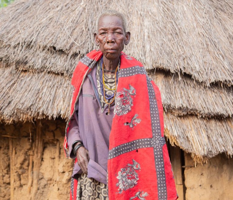 a Maasai elder and a visitor talking directly at the entrance to a manyatta homestead, warm afternoon light, Maasai Mara region grassland behind them