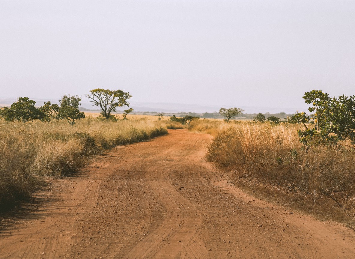 A red-dirt track winding through dry savanna grassland toward the horizon, the kind of overland road linking the Mara ecosystem to the Kenya-Tanzania border