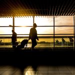 Travelers with luggage silhouetted against a departure lounge window overlooking the tarmac at golden hour, the kind of international terminal scene JKIA's new build aims to match