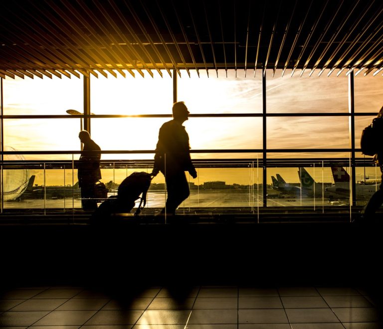 Travelers with luggage silhouetted against a departure lounge window overlooking the tarmac at golden hour, the kind of international terminal scene JKIA's new build aims to match