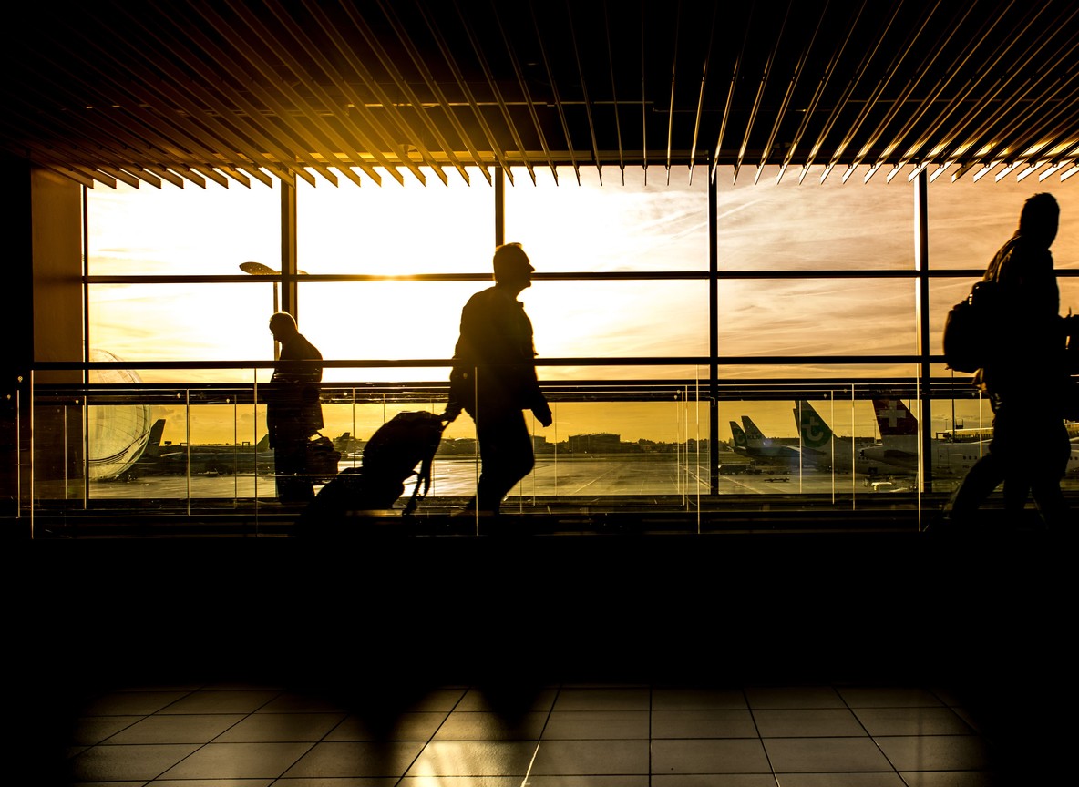 Travelers with luggage silhouetted against a departure lounge window overlooking the tarmac at golden hour, the kind of international terminal scene JKIA's new build aims to match