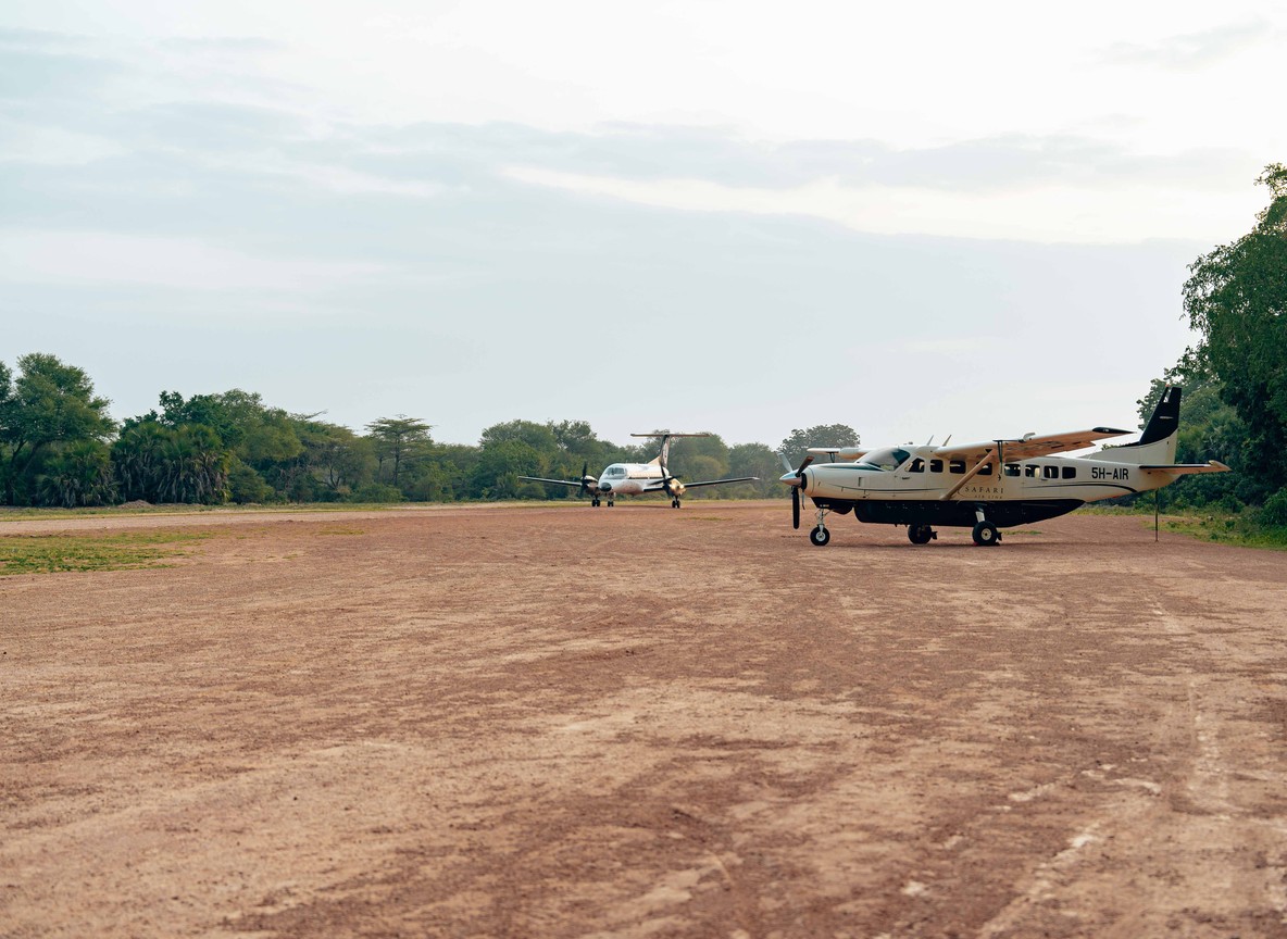 Small safari aircraft parked on a dirt airstrip with a safari vehicle waiting nearby