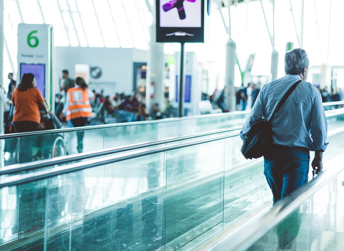 a traveler with a shoulder bag walking down an airport terminal walkway past gate signage