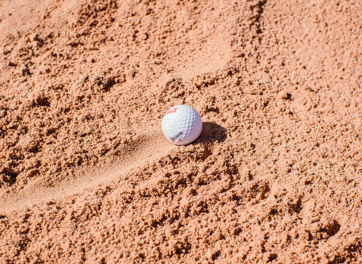 Close-up of a golf ball sitting in a sand bunker, illustrating a hazard travelers will meet on coastal fairways