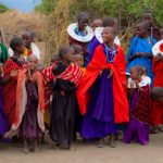 A joyful group of Maasai children and young women in traditional beaded collars and colourful shukas at a community gathering, the kind of village visit that pairs with a herder-led walking safari in Laikipia