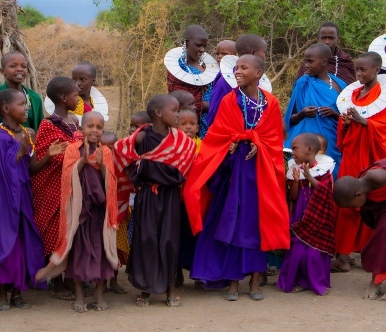 A joyful group of Maasai children and young women in traditional beaded collars and colourful shukas at a community gathering, the kind of village visit that pairs with a herder-led walking safari in Laikipia