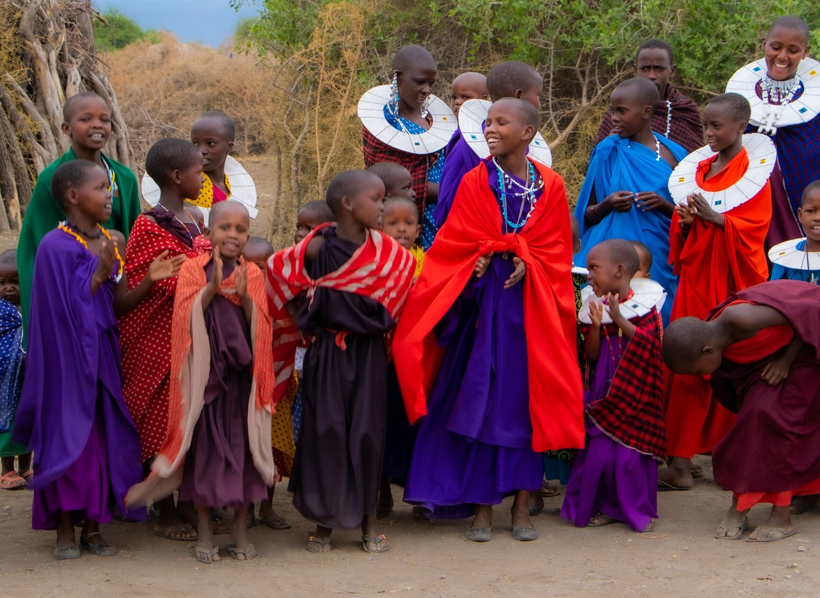 A joyful group of Maasai children and young women in traditional beaded collars and colourful shukas at a community gathering, the kind of village visit that pairs with a herder-led walking safari in Laikipia