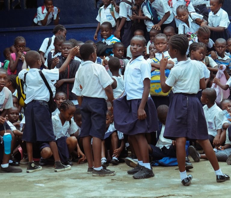 Kenyan primary school children seated outdoors listening to a teacher read a picture book, acacia trees and open Masai Mara grassland in the background