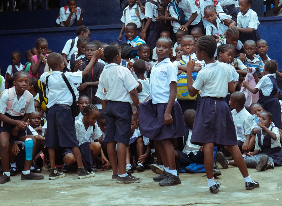 Kenyan primary school children seated outdoors listening to a teacher read a picture book, acacia trees and open Masai Mara grassland in the background