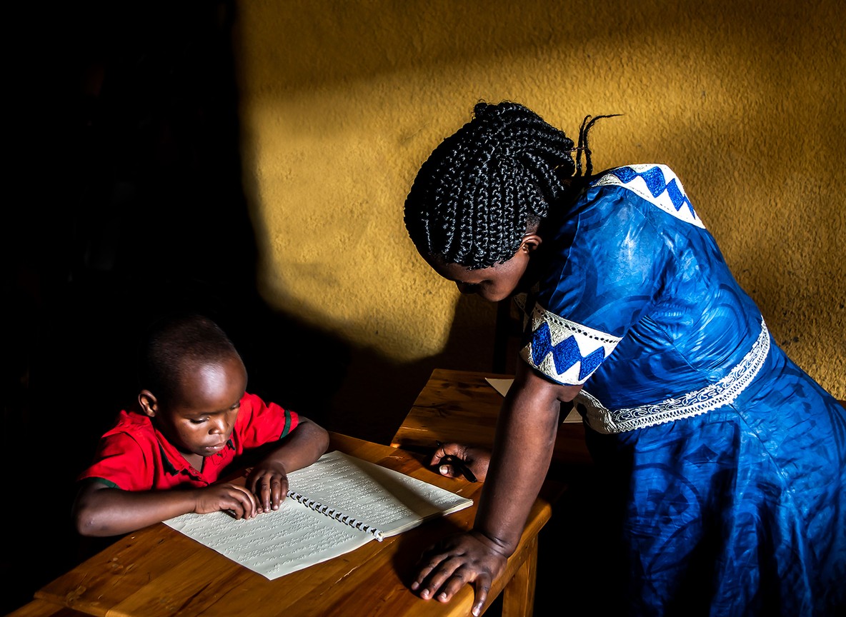 A young Eco-Mentor leading a small group discussion with schoolchildren outdoors near a Masai Mara village, notebooks and a picture book visible