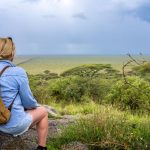 a solo woman traveler with a daypack sitting on a rock overlooking the open savanna, scanning the plains before her safari
