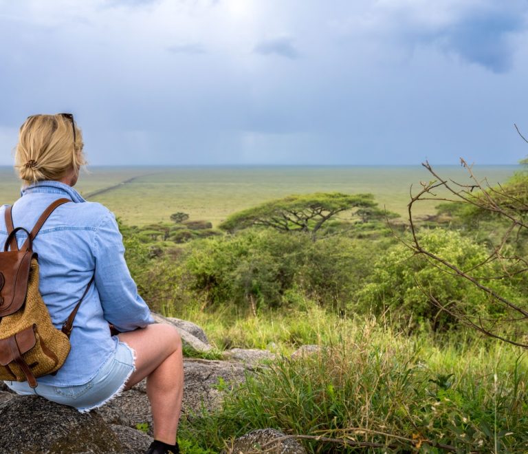 a solo woman traveler with a daypack sitting on a rock overlooking the open savanna, scanning the plains before her safari