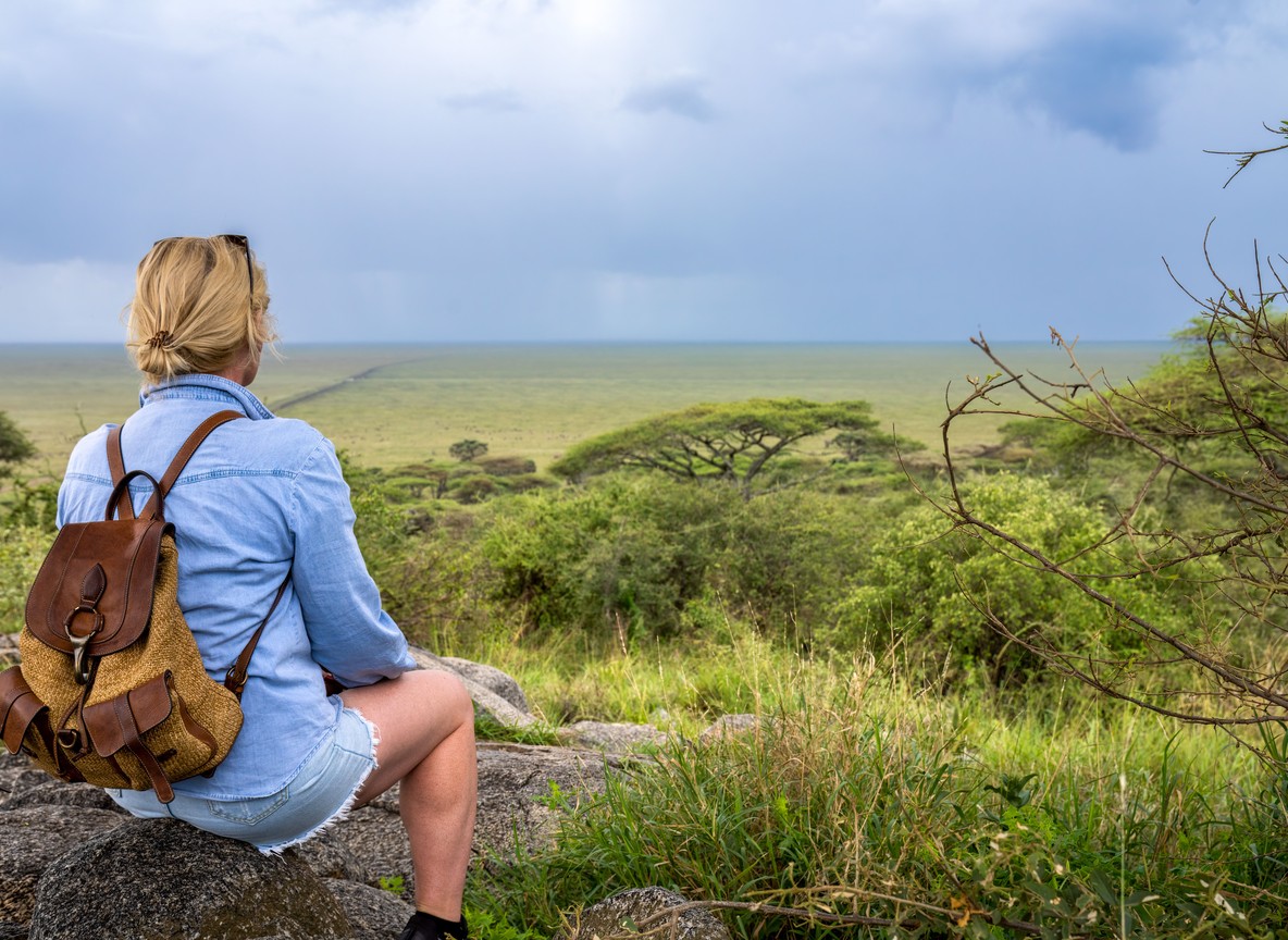 a solo woman traveler with a daypack sitting on a rock overlooking the open savanna, scanning the plains before her safari