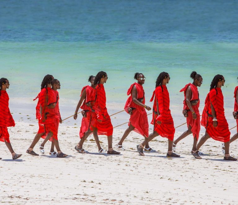 a line of newly graduated community scouts in field uniform standing at attention on dry red earth with the Chyulu Hills faint in the background