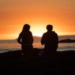 A couple standing together at a sunset viewpoint over the Maasai Mara plains, golden light across the grassland below, a quiet moment away from camp