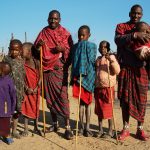 A mobile health outreach tent set up near a Maasai homestead with grassland and acacia trees in the background, safari vehicle parked nearby