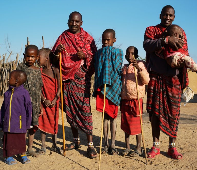 A mobile health outreach tent set up near a Maasai homestead with grassland and acacia trees in the background, safari vehicle parked nearby