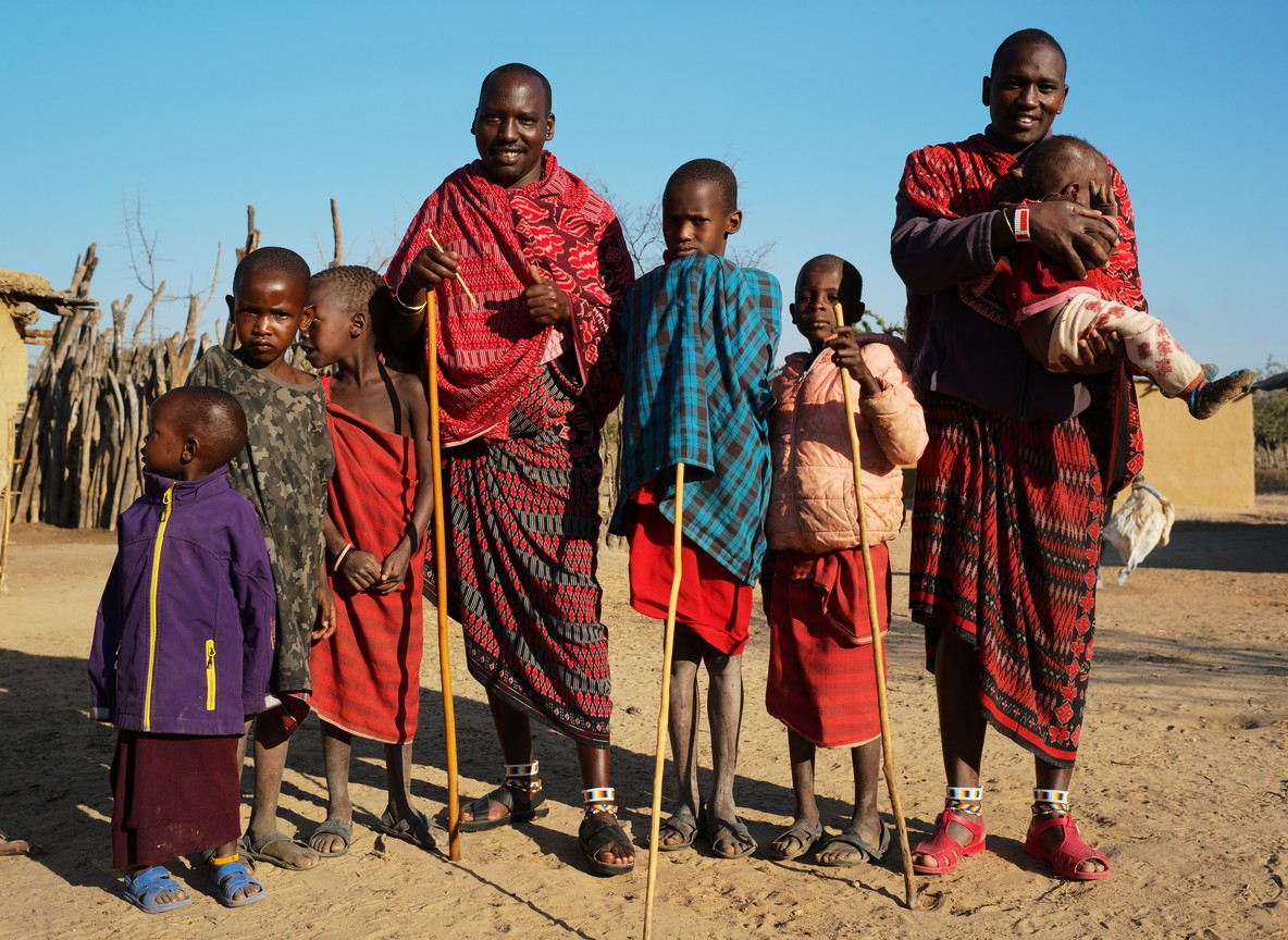 A mobile health outreach tent set up near a Maasai homestead with grassland and acacia trees in the background, safari vehicle parked nearby